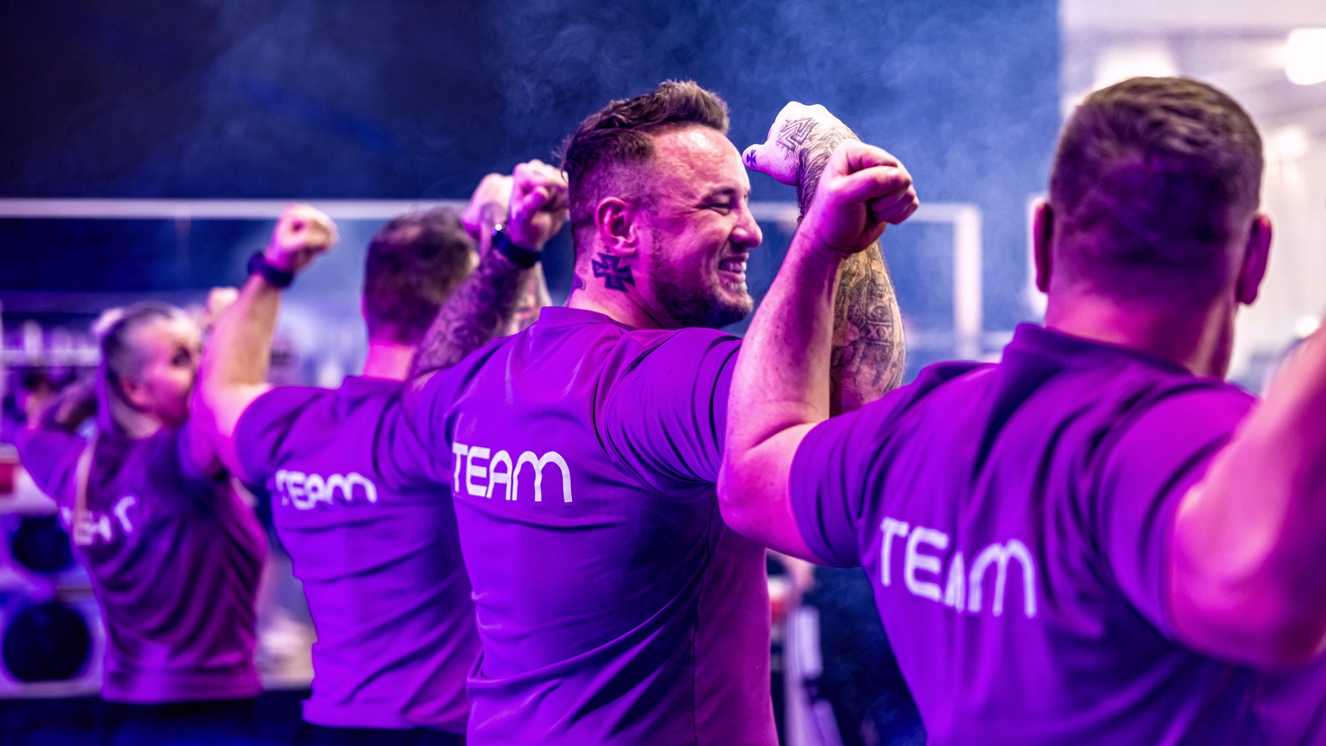FITfactory-Teammitglieder trainieren gemeinsam in violett beleuchteter Studioatmosphäre.